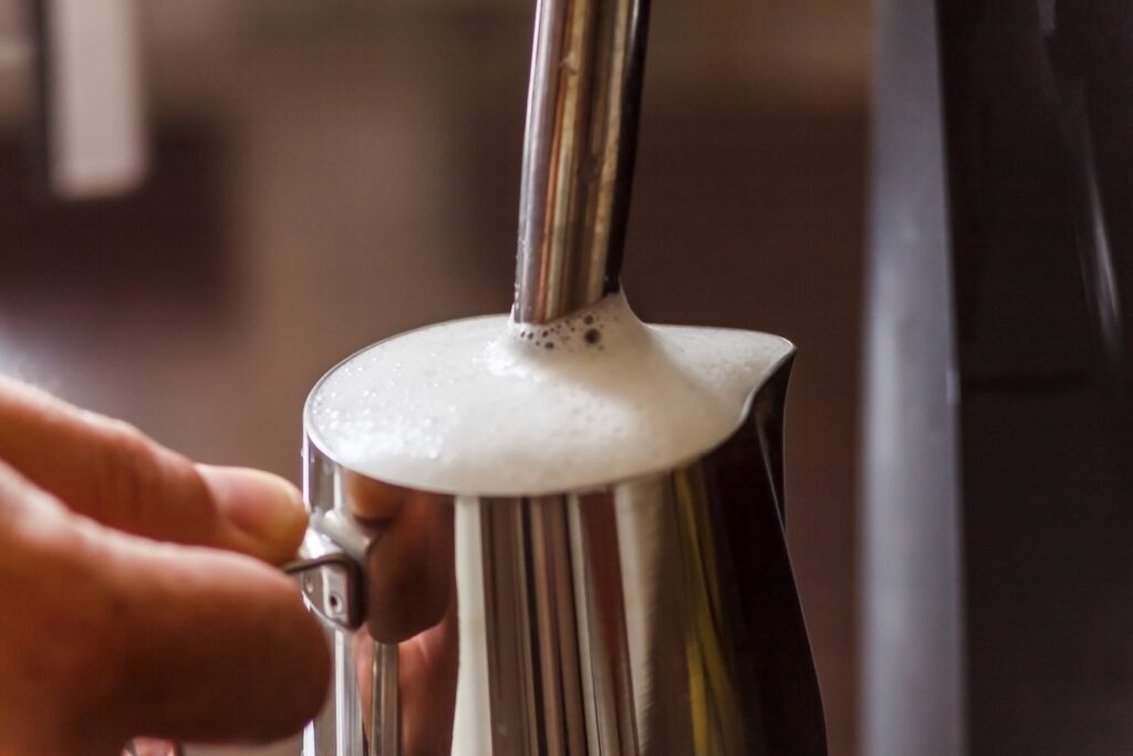 Close-up of a steam wand frothing milk in a stainless steel jug. The wand tip sits just below the surface, the aeration position that builds the foam, and a hand grips the jug by the handle to judge the temperature by feel. A layer of white microfoam has risen to the lip of the jug.