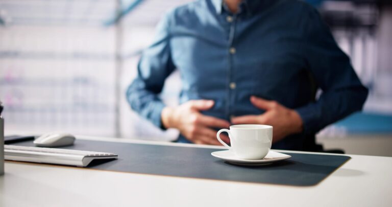 A man in a blue denim shirt at an office desk holding his stomach in discomfort, with a white coffee cup on a saucer in the foreground.