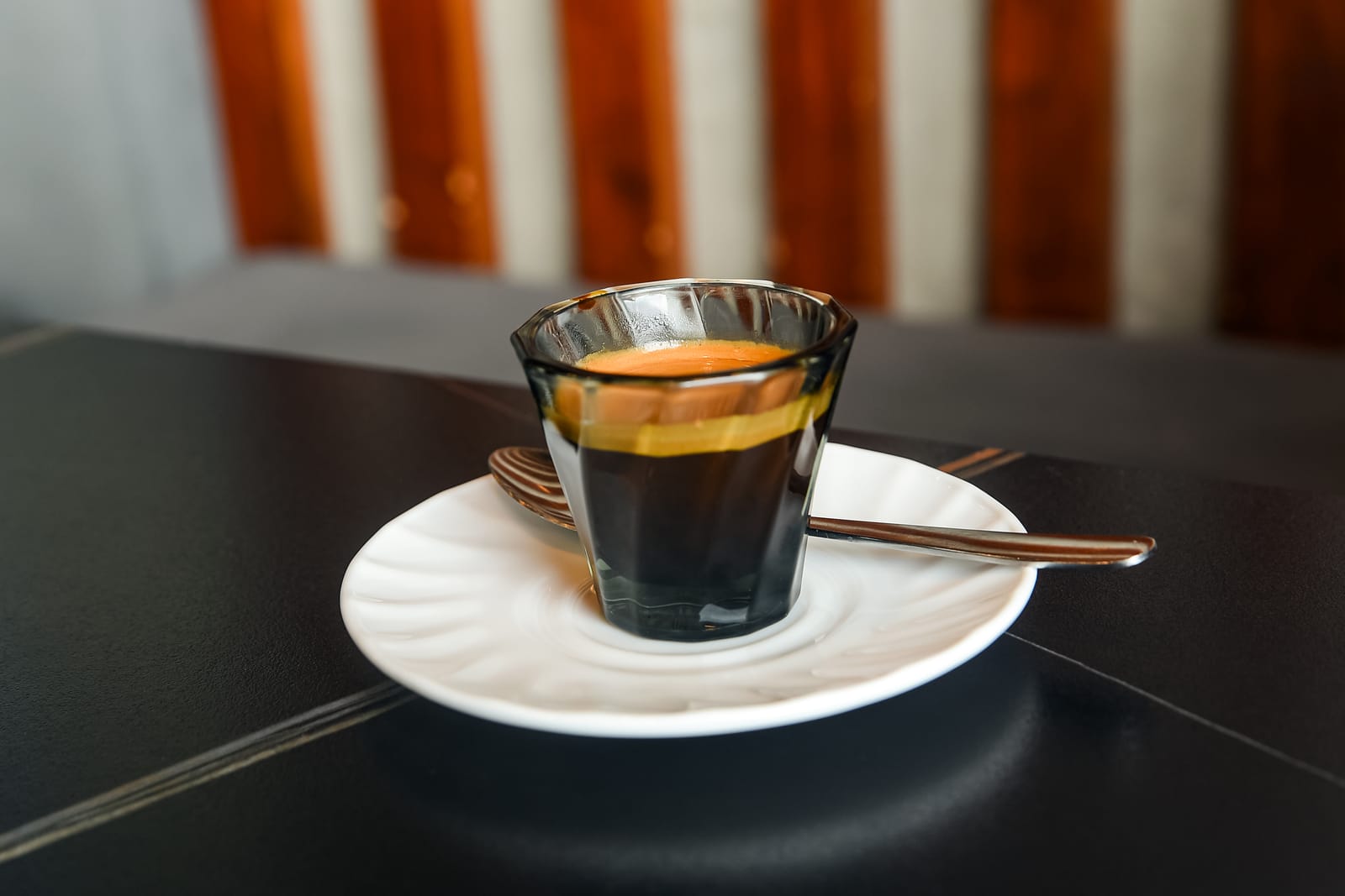 A single shot of espresso in a small smoked-glass tumbler, its orange crema sitting on top of the dark coffee, on a white saucer with a spoon.