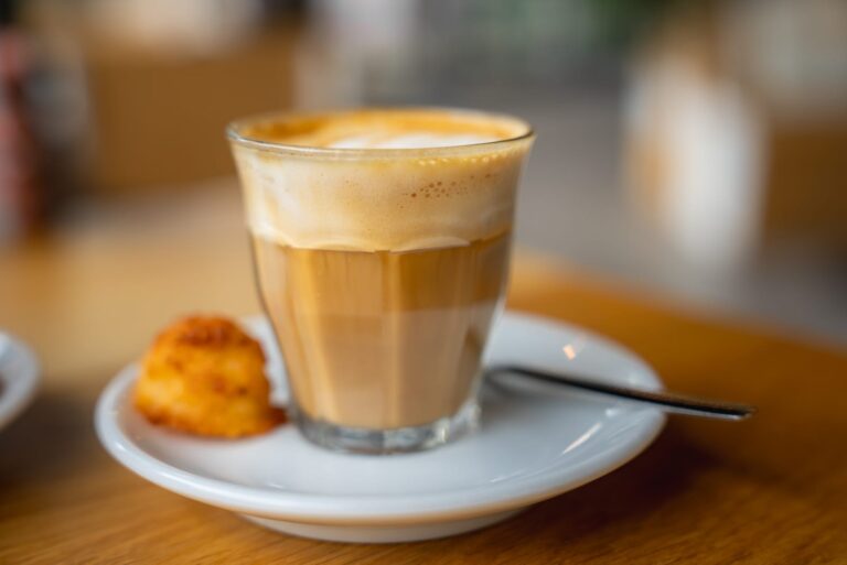 Latte macchiato in a small glass showing layers of milk, espresso and foam, on a saucer beside a small baked treat